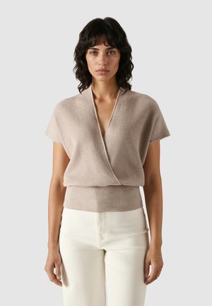 Mujer con cabello rizado y oscuro, vestida con un suéter cruzado de manga corta beige y pantalones blancos, de pie contra un fondo claro y liso.