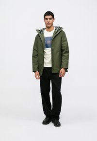 Casaco com capuz verde-oliva, com forro interior estampado, usado por cima de uma t-shirt gráfica clara e calças pretas de perna larga. Sapatos pretos.