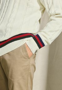 Fetter, weißer Kabelstrickpullover mit gerippten Bündchen und marineblauen, roten und grünen Streifen; kombiniert mit taupefarbenen Hosen.