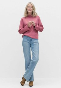 Rosa stickad tröja med hög krage, matchad med ljusblå utsvängda jeans och bruna ankelboots, mot en vit bakgrund.