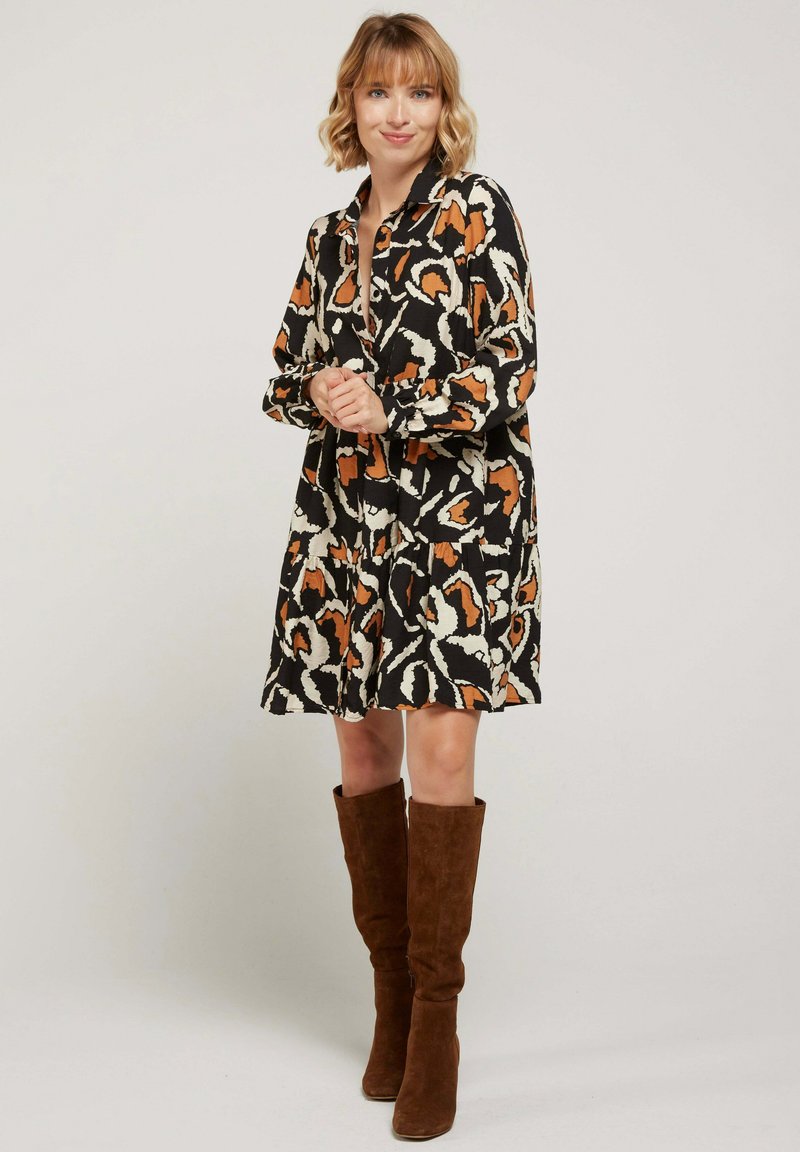 Robe noire avec un motif abstrait orange et crème, manches longues, bouton avant et une silhouette évasée. Bottes en daim marron montantes jusqu'aux genoux.