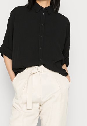 Vrouw draagt een losse zwarte blouse met knopen en opgerolde mouwen, ingestopt in een hooggetailleerde offwhite broek met stoffen riem.
