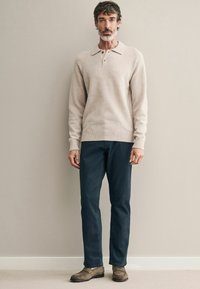 Suéter de lana beige con cuello y detalle de botones, combinado con jeans azul oscuro y zapatos de gamuza marrón. Textura suave, corte casual.