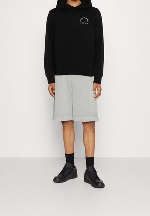 Sort hættetrøje med frontlomme og hvidt logo, kombineret med lysegrå shorts med sidelommer, båret med sorte sneakers og sokker.