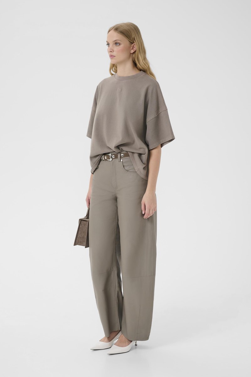 Femme portant un t-shirt taupe oversized, pantalon à jambes larges, talons blancs, tenant un petit sac à main marron, debout devant un fond uni.
