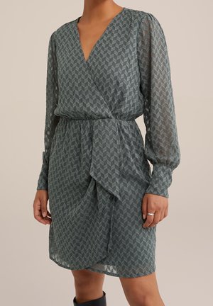 Kvinde iført knælængde, langærmet, tekstureret grå slå-om-kjole med subtilt geometrisk mønster og en talje med rynkede detaljer, stående med den ene hånd synlig.