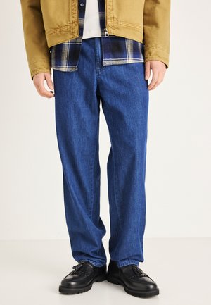 Dunkelblaue Jeans aus Denim mit geradem Bein und leichter Struktur. Kombiniert mit einem karrierten Hemd und einer hellbraunen Jacke, getragen mit schwarzen Schuhen.