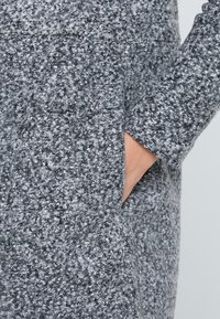 Szara, teksturowana tkanina ukazująca zbliżenie na kieszeń płaszcza. Materiał wydaje się miękki i puszysty, z połączeniem jasnych i ciemnych odcieni szarości.