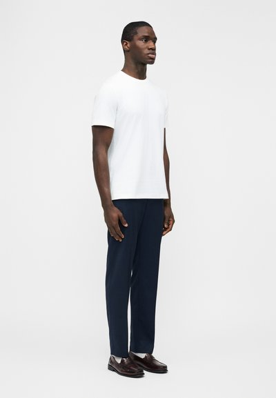 Άνδρας στέκεται φορώντας ένα απλό λευκό κοντομάνικο πουκάμισο, σκούρα μπλε παντελόνια, λευκές κάλτσες και σκούρα καφέ δερμάτινα παπούτσια μπροστά σε λευκό φόντο.