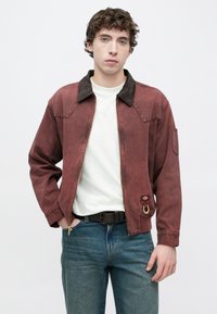 Κόκκινο-καφέ μπουφάν με καφέ γιακά, εμπρόσθιο φερμουάρ και πλαϊνή τσέπη. Διαθέτει ένα μικρό λογότυπο και μια μεταλλική διακόσμηση σε σχήμα δακτυλίου στο εμπρόσθιο μέρος.