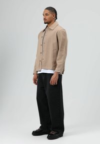 Beige katoenen jas met knoopsluiting, twee zijzakken, gecombineerd met zwarte jeans en chunky zwarte schoenen met roze accenten.