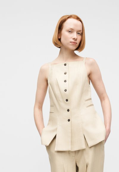 Γυναίκα με κοντά κόκκινα μαλλιά, που φοράει μπεζ αμάνικο τοπ με κουμπιά και παντελόνι που ταιριάζει, στέκεται με τα χέρια στις τσέπες μπροστά σε ένα απλό φόντο.