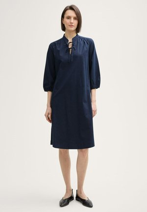 Rochie bleumarin, până la genunchi, cu guler înalt, mâneci half-sleeve și un decolteu cu șireturi. Țesătură netedă cu drapare moale și deschideri laterale.