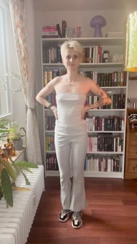 Person with short blonde hair, white strapless top, white pants, and sneakers standing with hands on hips in front of bookshelf.