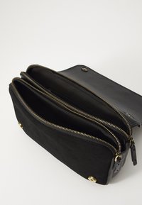 Svart mockaväska med dragkedja-design, som har tre fack och detaljer i guldfärg. Slät textur med knappstängning.