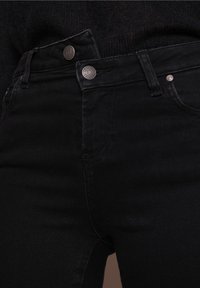 Czarne jeansy z denimu mają dopasowany fason z dwoma srebrnymi metalowymi guzikami na pasku oraz minimalistyczne detale przeszyć.