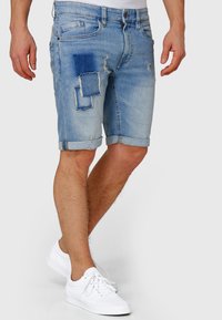 Pantaloni scurți din denim albastru deschis cu margini zdrențuite, cu detalii tip patch-uri și croială slim. Asortați cu adidași albi.