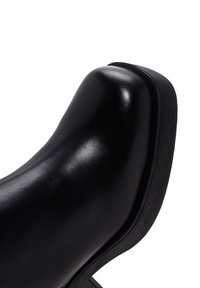 Stivale in pelle nera lucida con punta quadrata arrotondata e suola spessa in gomma nera su sfondo bianco.
