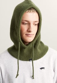 Zelená žebrovaná pletená kapuce s tkani�čkami, s širokým límcem, nošená přes šedou mikinu s malým černým logem skateboardu.