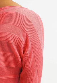 Lekki sweter koralowy z paskowym wzorem, z naprzemiennymi gładkimi i prążkowanymi sekcjami, prezentujący luźny krój oraz miękki materiał.