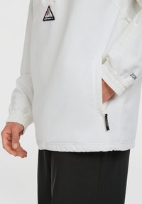 Witte windbreaker jas met elastische manchetten, een zijzak met een zwarte rits en een driehoekige logo patch op de borst.