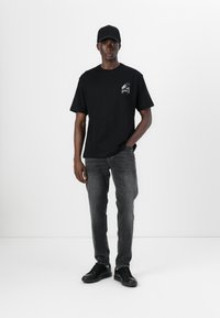 Black cotton t-shirt with short sleeves, round neckline, and a small graphic on the chest. Paired with gray slim-fit jeans and black sneakers.