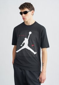 Μαύρο βαμβακερό T-shirt με μεγάλο λευκό σιλουέτα Air Jordan και κόκκινο κείμενο σε μοτίβο πλέγματος. Συνδυάστε το με σκούρο αθλητικό παντελόνι και γυαλιά ηλίου.