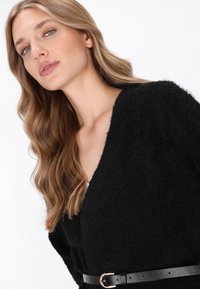 Zwarte fuzzy cardigan met een diepe V-halslijn, voorzien van een zwarte riem met een gouden gespaccent aan de taille. Zachte textuur.