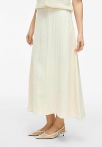 Jupe maxi couleur crème avec une texture froncée, un design fluide et une longueur jusqu'aux chevilles. Associée à des escarpins à talons ouverts rose pâle.