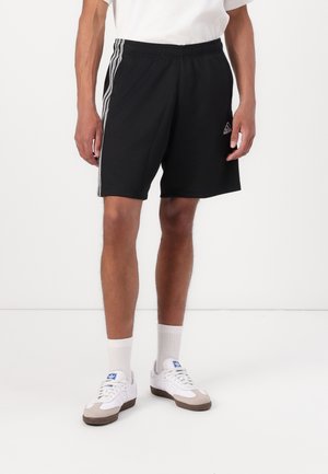 Man draagt zwarte Adidas-short met witte drie strepen aan de zijkanten, witte sokken en witte Adidas-sneakers, staand op een witte vloer.