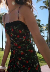 Svart blommig klänning med spetskant, prydd med röda jordgubbar och gröna blad. Tunna axelband och mjukt tyg. Miljön inkluderar palmer.