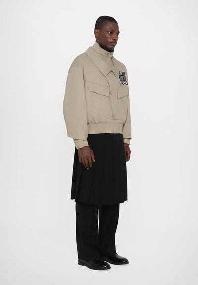 Beige Cropped-Jacke mit großen Taschen und hohem Kragen, kombiniert mit einem schwarzen Faltenrock und schlanken schwarzen Schuhen.