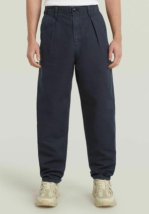 Homme portant un pantalon plissé bleu marine foncé et des baskets beiges épaisses, debout sur un fond neutre.