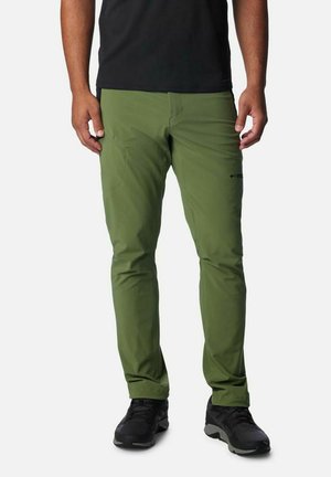 Mann trägt olivgrüne, schmal geschnittene Hose und schwarze Turnschuhe, steht vor einem schlichten weißen Hintergrund.