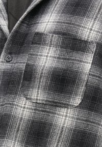 Καρό πουκάμισο σε ασπρόμαυρο με υφή στο ύφασμα, διαθέτει κλείσιμο με κουμπιά και τσέπη στο στήθος. Ο σχεδιασμός περιλαμβάνει ομοιόμορφα μοτίβα καρό.