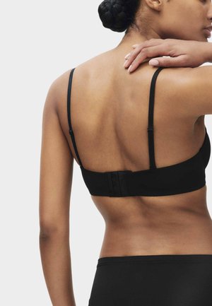 Frau mit geflochtenem Haar, die einen schwarzen verstellbaren BH und passende Unterteile trägt, von hinten gezeigt, eine Hand ruht auf der Schulter.