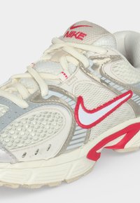 Λευκό και γκρι αθλητικό παπούτσι Nike με κόκκινες λεπτομέρειες, πλέγματα και λευκά κορδόνια, ορατό σε ανοιχτό φόντο.