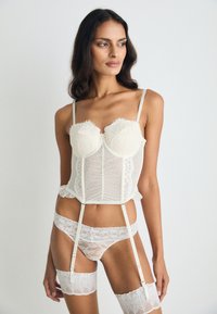 Κρεμ κορσέ με δαντελένια τελειώματα, διάφανο ύφασμα με πουά, ρυθμιζόμενους ιμάντες και αντίστοιχα δαντελωτά εσώρουχα με καλσόν. Λείο υλικό σε όλη την επιφάνεια.