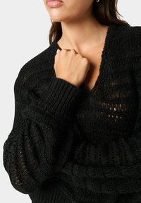 Schwarzer gestrickter Pullover mit V-Ausschnitt, der ein strukturiertes Muster und gerippte Ärmel aufweist und eine lockere, übergroße Passform betont.