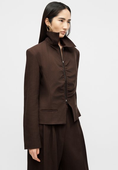 Γυναίκα με μακριά σκούρα μαλλιά, φορώντας καφέ μπουφάν με φερμουάρ και ψηλό γιακά, καθώς και παντελόνι φαρδύ που ταιριάζει, κοιτάζοντας προς τα δεξιά.