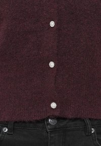 Pull en tricot bordeaux avec une finition texturée, comportant quatre boutons décoratifs en perles sur le devant et un ourlet brut.