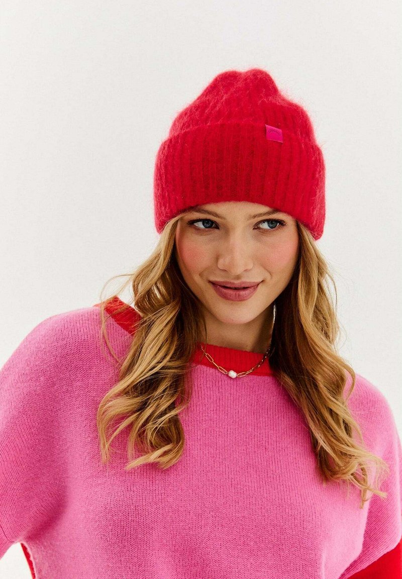 Červená pletená čiapka s rebrovanou textúrou a malou ružovou značkou. Nosí sa s ružovým svetrom s kontrastným červeným výstrihom.