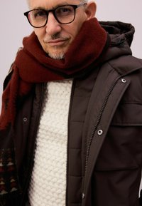 Gepolsterte dunkelbraune Jacke mit Reißverschlüssen, cremefarbener Strickpullover in Zopfstrick und ein roter Wollschal mit geometrischem Muster.