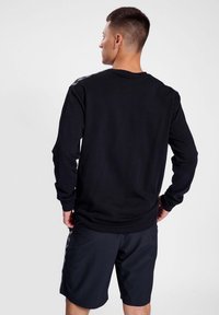 Maglietta nera a maniche lunghe con colletto rotondo e polsini a costine, abbinata a pantaloni corti scuri; texture liscia e design minimalista.
