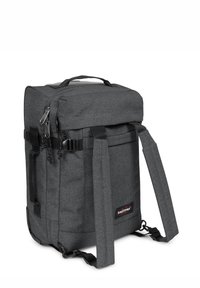 Mochila de tela gris con forma rectangular, asa superior, correas ajustables, cremalleras y logo de Eastpak. Presenta una superficie texturizada.