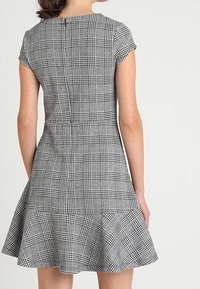 Robe à carreaux gris avec des manches courtes, un ourlet évasé et un corsage ajusté. Présente un tissu texturé avec un motif visible de carrés et de lignes.