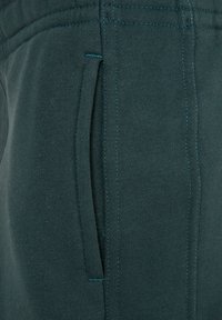 Dunkelgrüne Sweatpants mit einer weichen Textur, ausgestattet mit einer Seitentasche und kontrastierenden Nähten. Der Stoff wirkt bequem und flexibel.