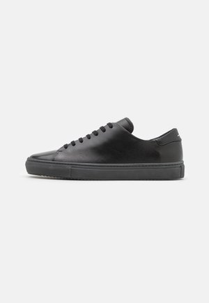 Zapatilla deportiva baja de cuero negro con cordones negros y suela de goma negra, mostrada de perfil sobre un fondo claro y liso.