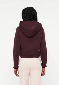 Orezan hoodie v burgundski barvi iz tkanine z žepom za kenguru na sprednji strani in velikim usnjem, v kombinaciji z teksturiranimi svetlimi hlačami. Brez vidnih oznak.