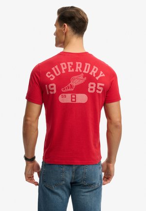 Mężczyzna noszący czerwoną koszulkę z napisem "SUPERDRY 1985" i grafiką skrzydlatej sandały, w niebieskich dżinsach, z tyłem do kamery.